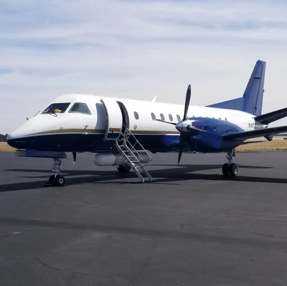 A small twin-engine propeller aircraft is parked on a tarmac under a partly cloudy sky. The plane has a white and blue color scheme, and a set of metal stairs is positioned at the open passenger door on the left side.