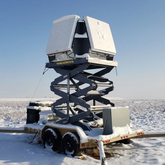 Four large square phased arrays on a scissor lift tower that is raised. The entire system sits on a trailer that is placed in the middle of a large snowy field. The tower and phased arrays are covered in snow, and the sky is perfectly blue.