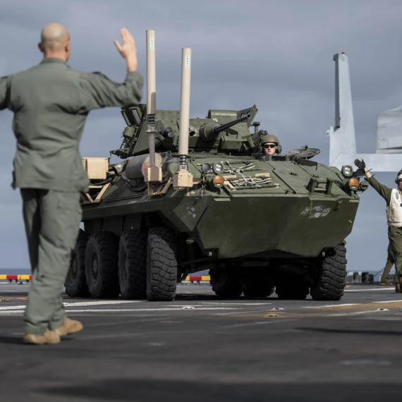 A military armored vehicle is being guided by personnel on the deck of a naval ship. One person in uniform stands with arms raised, directing the vehicle, while others observe. The armored vehicle is green and heavily equipped, and the setting appears to be during daytime under a cloudy sky.