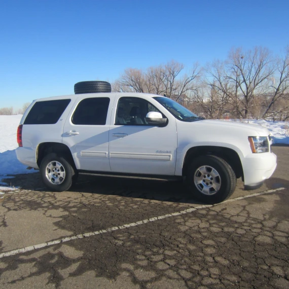 A white SUV pictured from the side with a tire attached to the top of the vehicle. It is resting on a cleared parking lot with snow in the background on a perfectly sunny day.