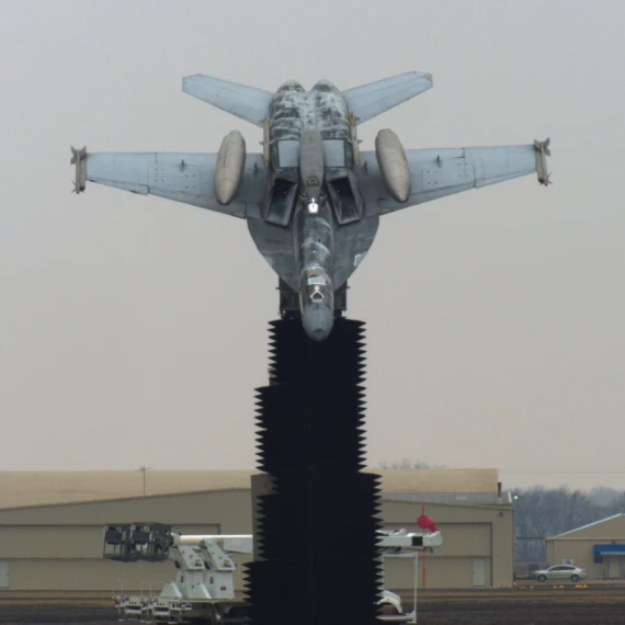 A fighter jet is mounted vertically on a tall black pedestal as a static display at an airfield. The aircraft is angled nose-down, with its landing gear retracted and wings outstretched. In the background, hangars and other airport structures are visible under an overcast sky.