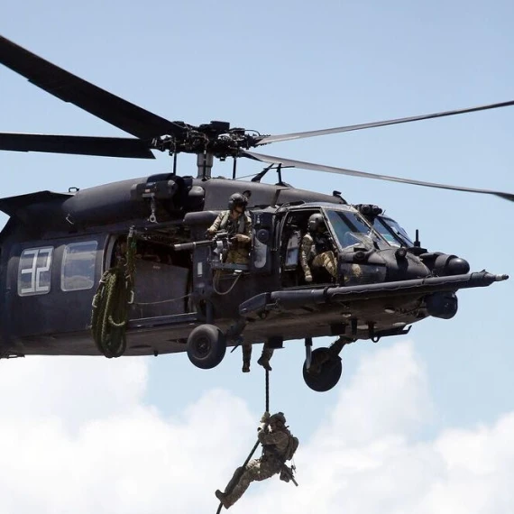 A military helicopter hovers in the air with armed soldiers aboard. One soldier is rappelling down a rope from the open side door of the helicopter, while others watch and assist. The sky is mostly clear with a few clouds in the background.