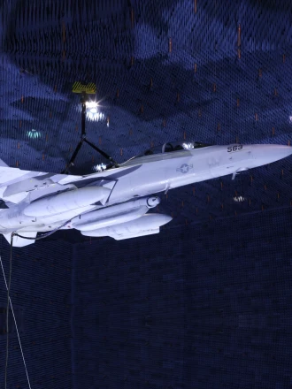 A Growler jet is shown suspended in a large anechoic chamber with three large pod-looking payloads mounted to it's belly. FIRST RF's NGJ-LB (Next Generation Jammer - Low Band) is the center pod on the belly of the aircraft.