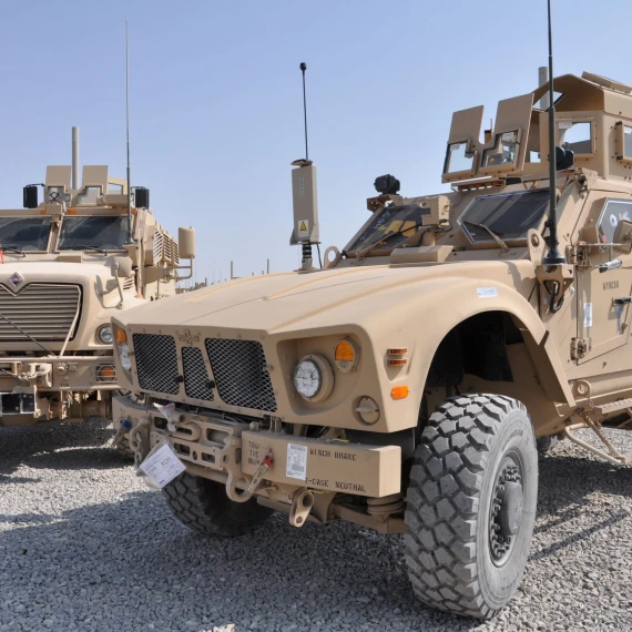 A close-up image of the front right-view of a M-ATV Army vehicle equipped with an assortment of antennas. There is an additional army vehicle in the background on the left side of the image. Both vehicles sit on a gravel ground and the sky is completely clear.