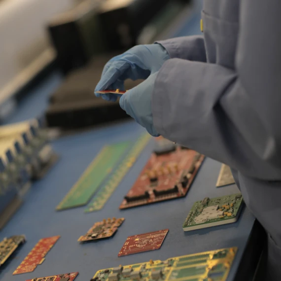 A blue table of a handful of printed circuit boards is shown. On the right, the torso of a technician, wearing a blue smock and blue plastic gloves, is shown holding one of the PCBs.